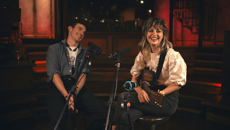 Dónal Finn and Anaïs Mitchell sit in front of two microphones laughing. Anaïs is holding an acoustic guitar.